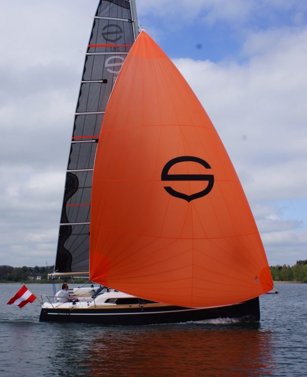 Eine Sunbeam 28.1 Segelyacht mit einem auffälligen orangefarbenen Gennaker, der das schwarze Sunbeam-Logo trägt, segelt auf einem See.
